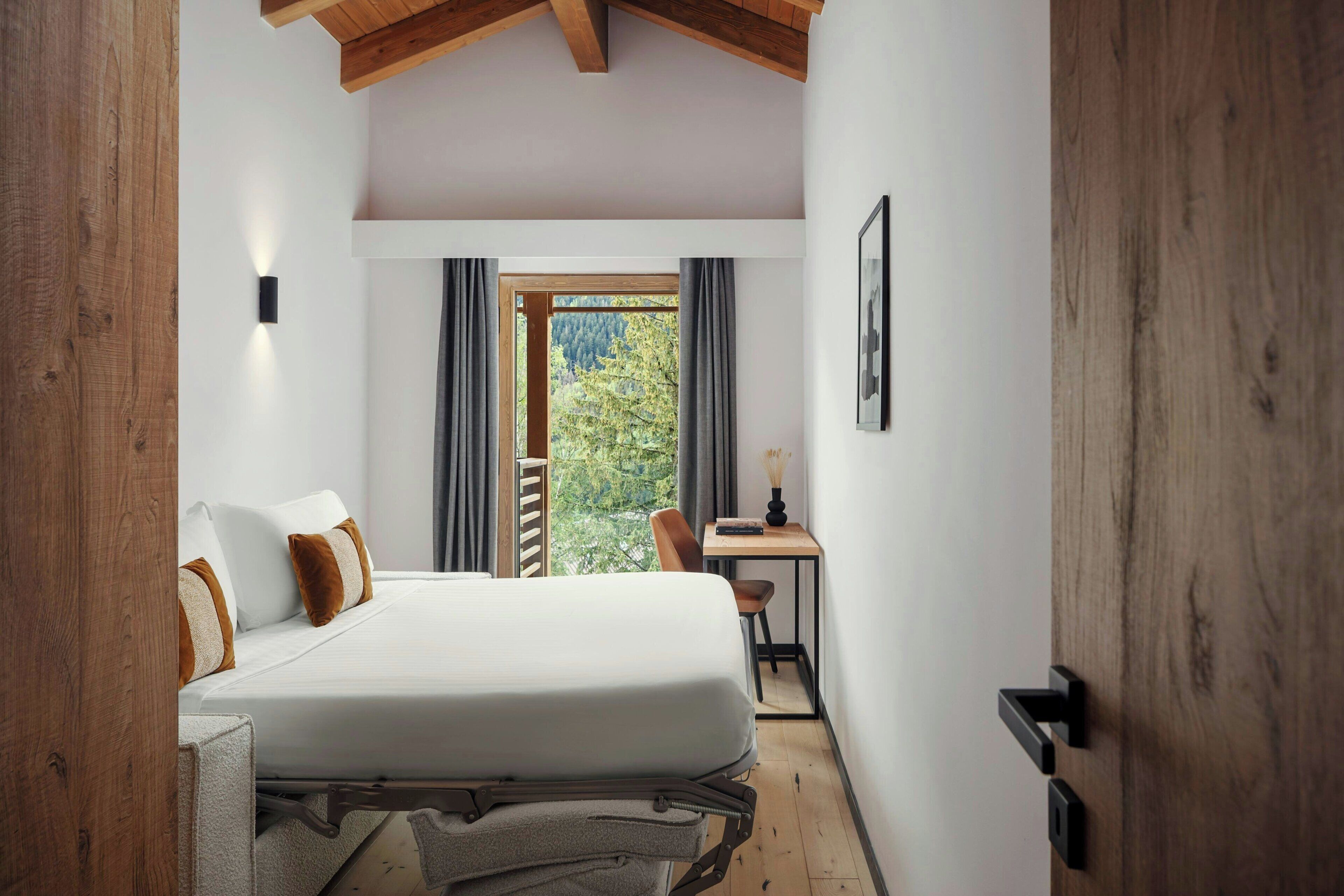 A bedroom with a bed, a desk, and a view of trees through a window.