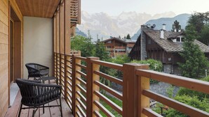 Un balcón de madera con dos sillas de mimbre y una mesa, con vistas a un paisaje montañoso y edificios tradicionales.