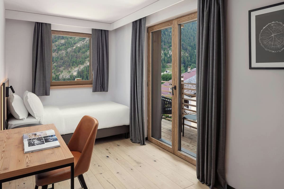 Ein modernes Schlafzimmer mit einem Bett, einem Holztisch mit einer Zeitschrift, einem Sessel und Blick auf eine gebirgige Landschaft durch ein großes Fenster.