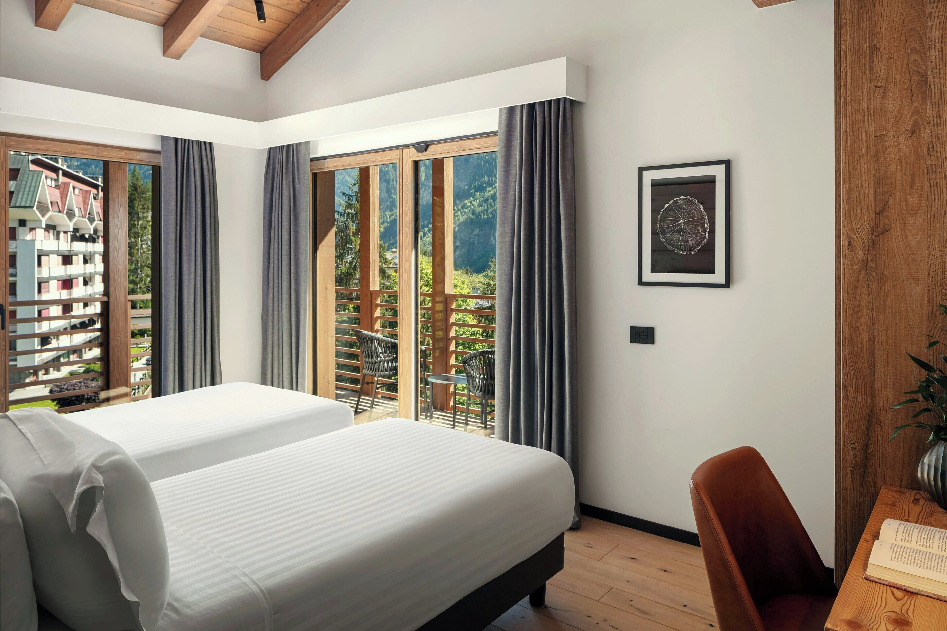 Ein Schlafzimmer mit zwei Betten, Holzboden, einem großen Fenster mit Vorhängen, einem gerahmten Bild an der Wand und einem Holztisch mit einem Buch.