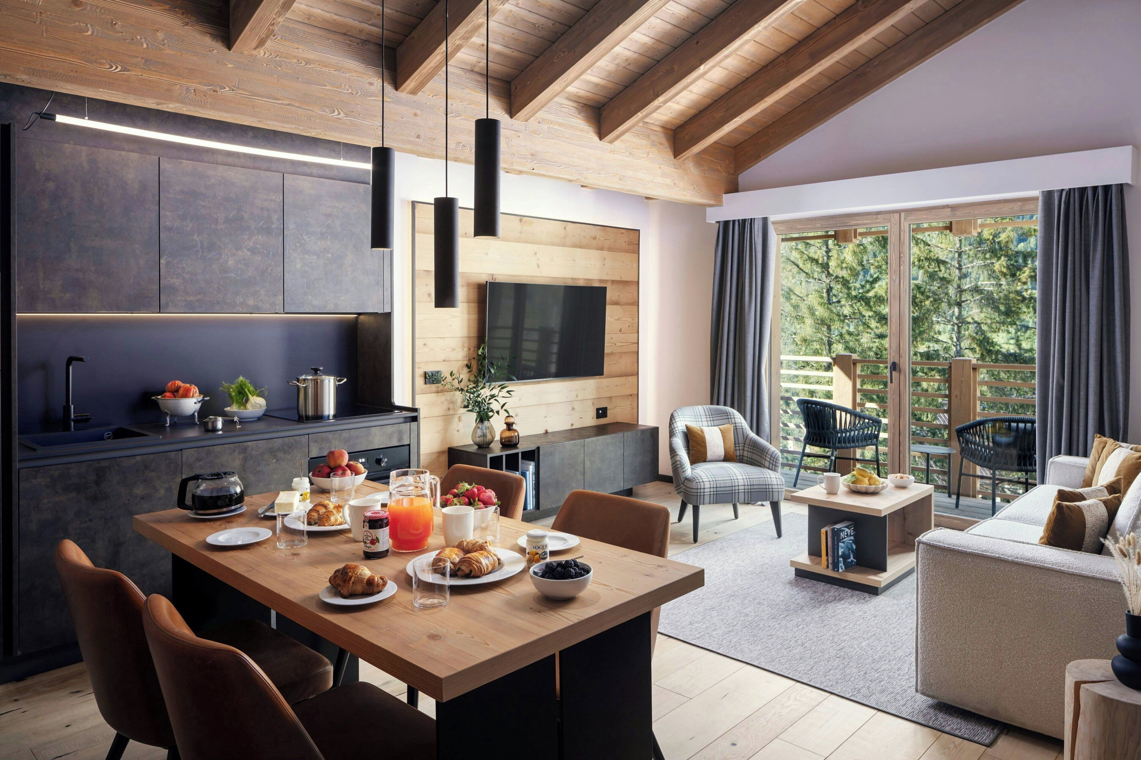 Una cocina moderna con comedor, vigas de madera en el techo y un amplio ventanal con vistas a un bosque.