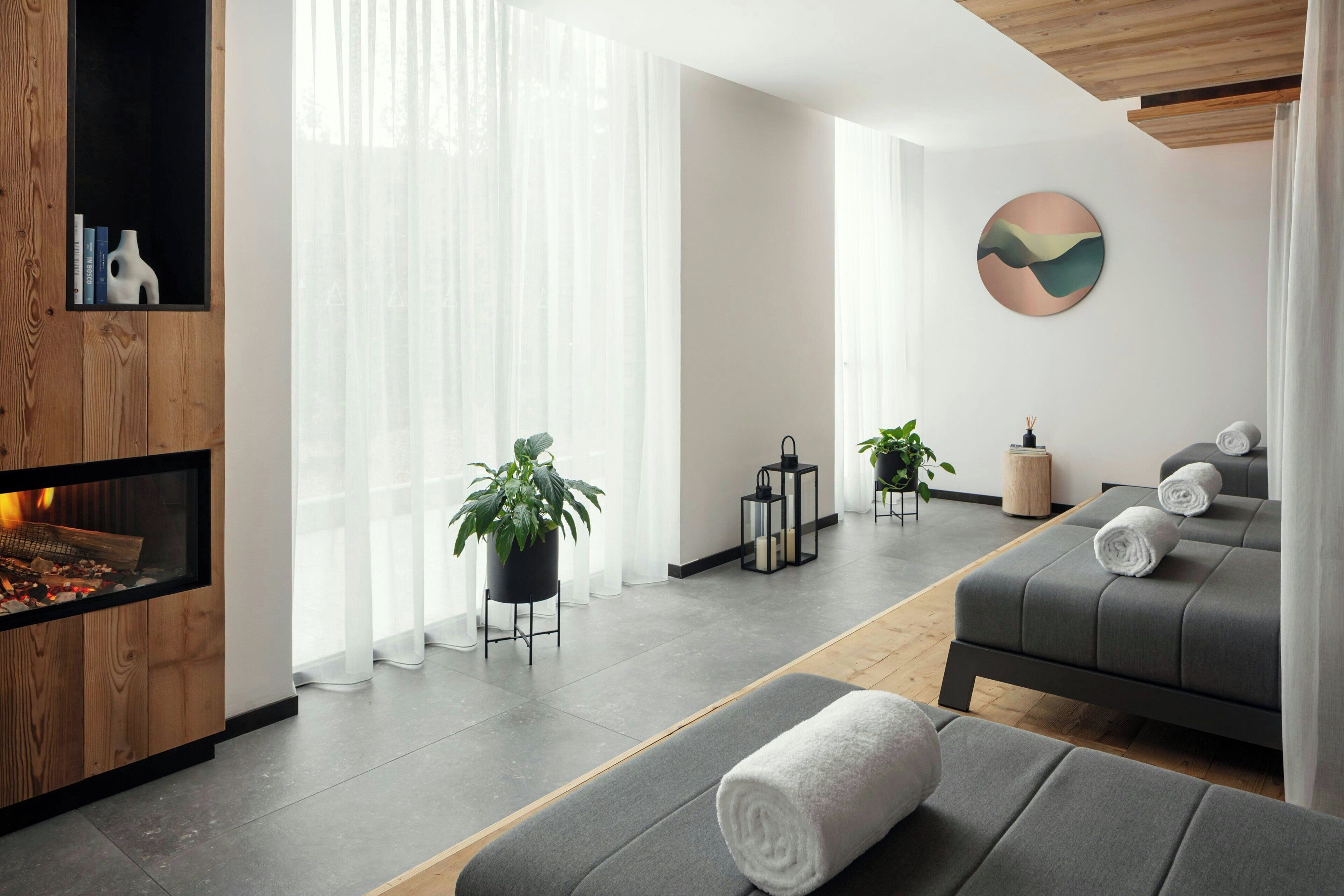 Una sala de estar moderna con chimenea, sofás grises, un ventanal con cortinas translúcidas y una pieza de arte circular en la pared.