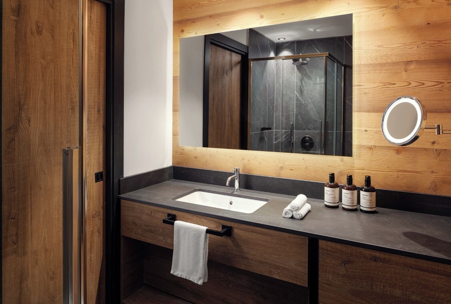 Ein modernes Badezimmer mit einem Holzwaschtisch, einem großen Spiegel, einer Dusche und drei Shampooflaschen.