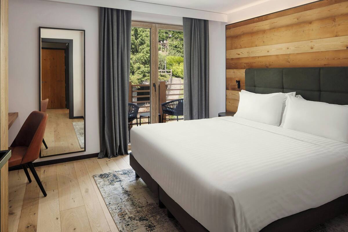Ein modernes Hotelzimmer mit einem großen Bett, einem Schreibtisch mit Stuhl und einem Balkon mit Tisch und Stühlen.
