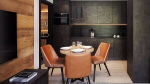 Un comedor moderno con una mesa redonda para cuatro, dos sillas de cuero marrón, una pared decorativa de madera oscura y una cocina integrada con muebles oscuros y una campana extractora de acero inoxidable.