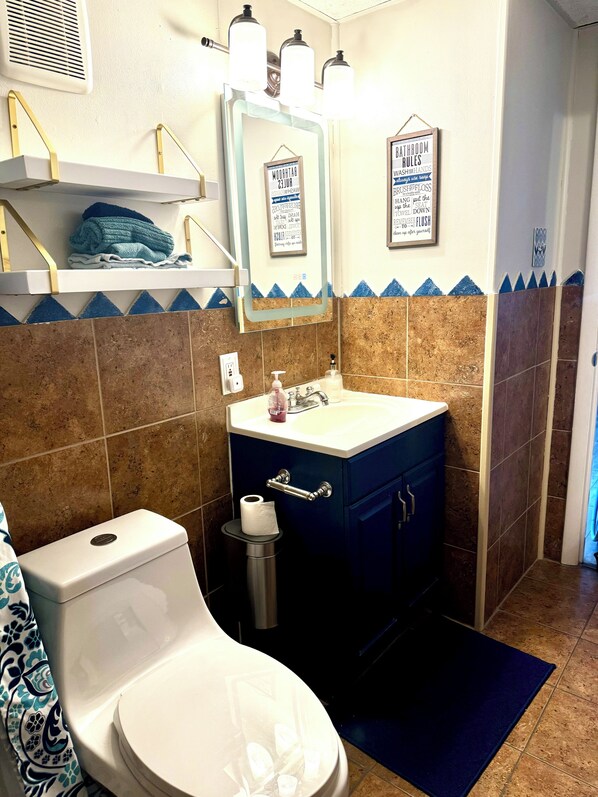 A bathroom with a toilet, sink, and mirror. There are towels on a shelf, a trash can, and a framed sign on the wall.