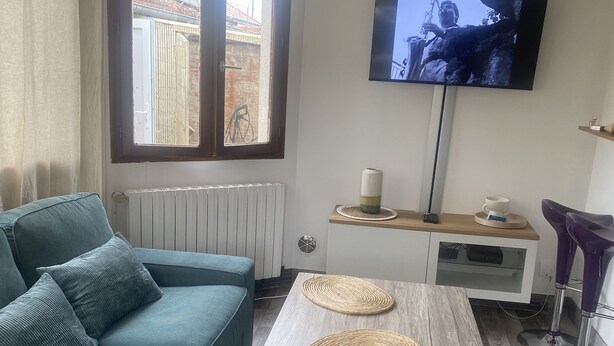 A living room with a teal sofa, a wooden coffee table with two woven placemats, a television mounted on the wall, and a window with a view of a building outside. - Quiet house in Vernon (Vernon)