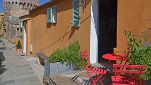 A narrow street with a building featuring a stone tower, a small garden, and a red table and chairs outside. - LATU CORSU – Cosy studio for 2 (ERSA)
