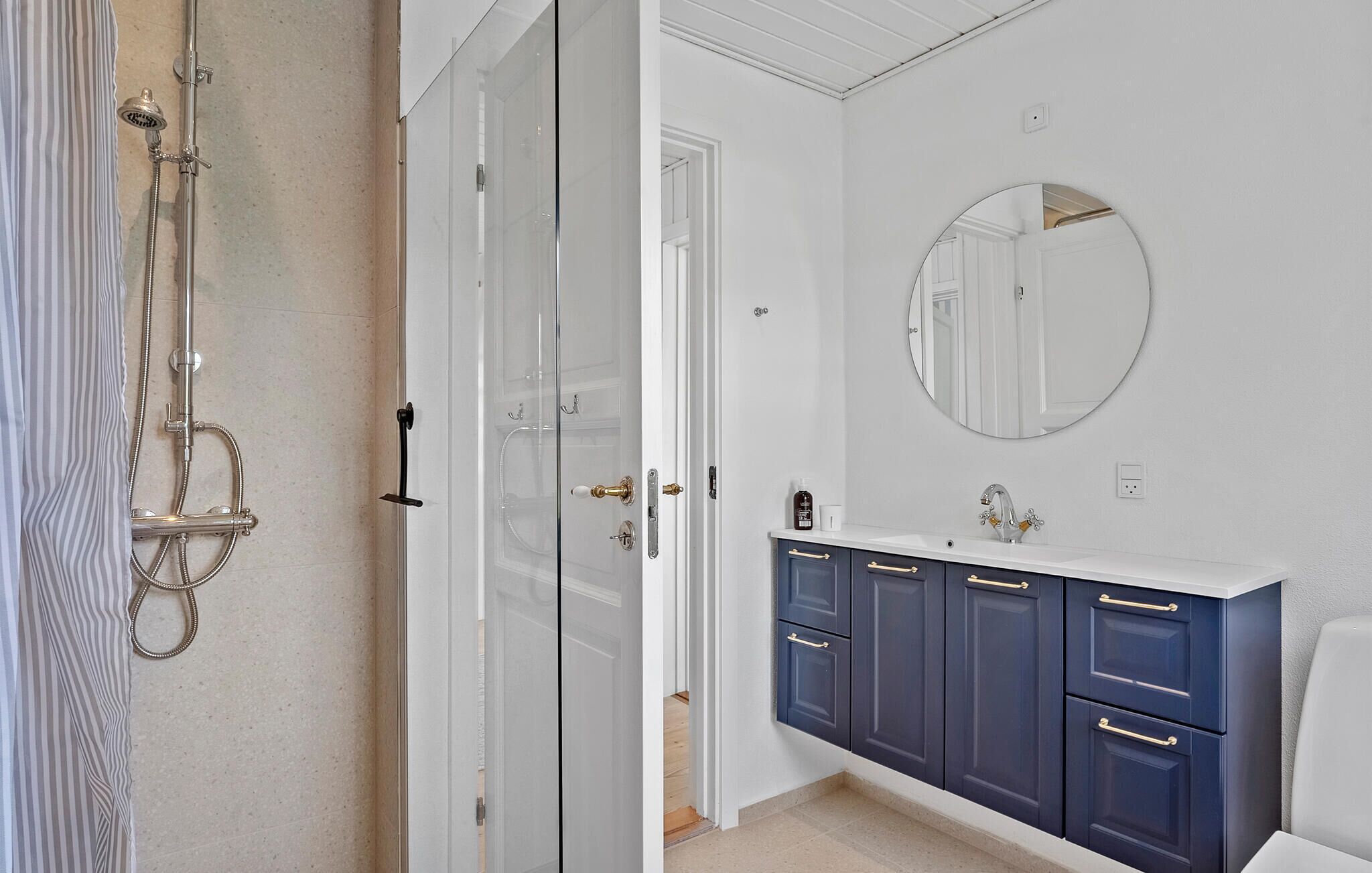 Een badkamer met een douche met glazen wand, een wit wastafelmeubel met donkerblauwe kastjes en een ronde spiegel aan de muur.