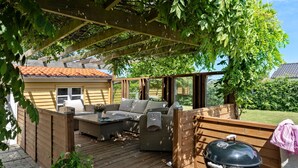 Et overdækket udendørs siddeområde med grill og et hus i baggrunden.