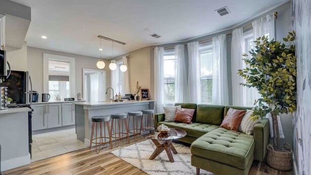 A modern kitchen with a central island, bar stools, and a green sofa in a living area with large windows and a plant. - SunSuite - 6 Min. Walk to Trains - Summer Special! (Jersey City)