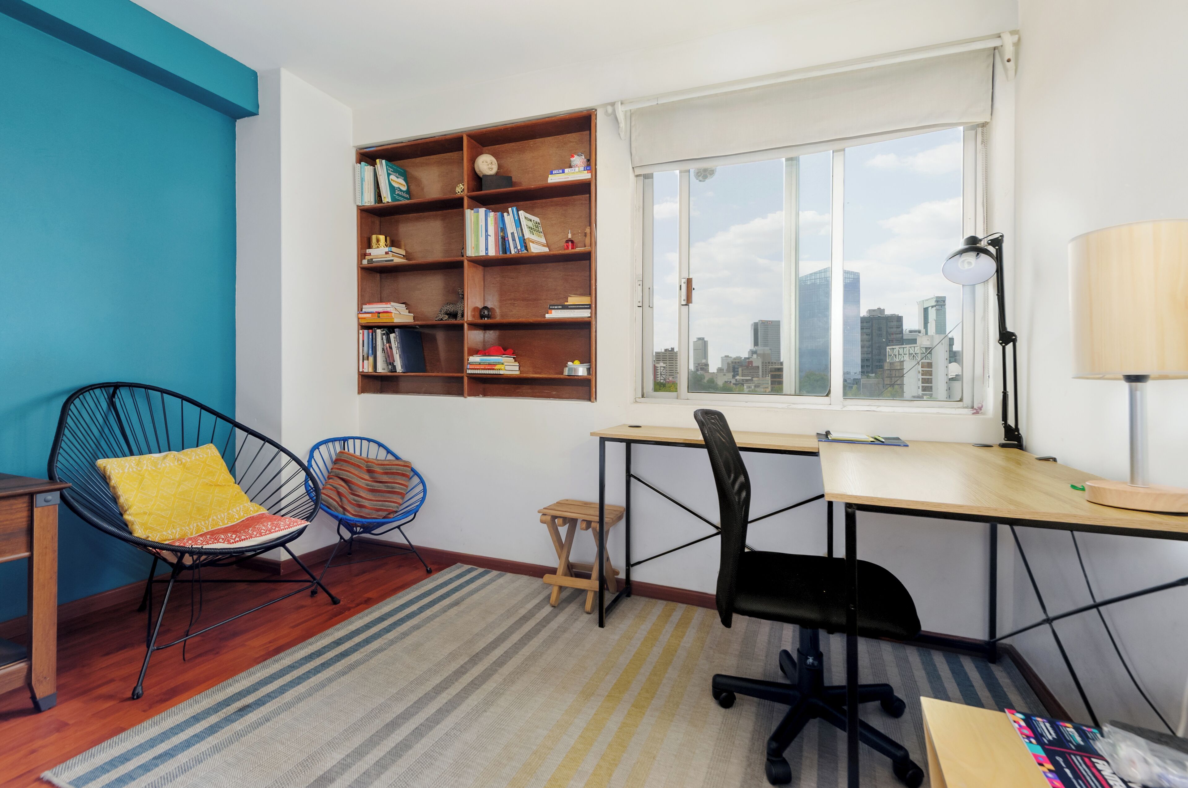 A room with a desk, two chairs, a bookshelf, a window with a city view, and a lamp.