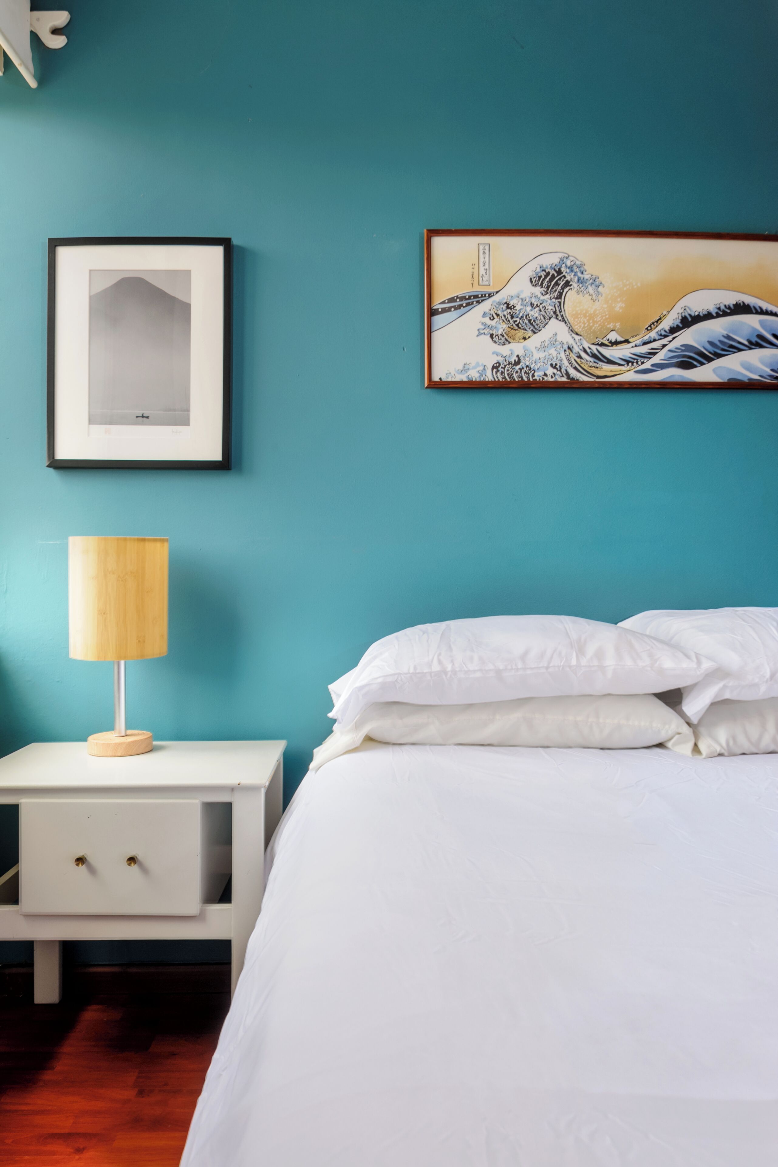 A bedroom with a bed, a nightstand with a lamp, and two framed artworks on the wall.