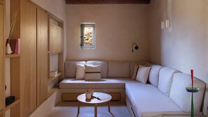 A cozy room with a wooden ceiling, a built-in bench with cushions, a small table with a book and a cup, and a window with a view of a rocky landscape. - Mousti's Cell Traditional Cycladic Farmhouse (Serifos Island)