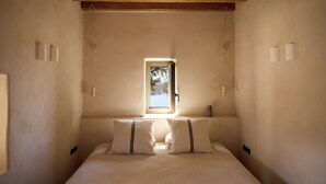 A bedroom with a bed, two bedside tables, a window, and wooden beams on the ceiling. - Mousti's Cell Traditional Cycladic Farmhouse (Serifos Island)