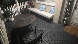 Een terras met een ronde tafel en stoelen, een witte hekwerk, een bank met kussens, en een 'WELCOME' bord.