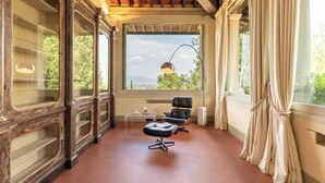 A room with a large window, a black leather chair, a small table with a lamp, and a view of greenery outside. - Villa Loretino - Ultimate Luxury in Florence (Comeana)