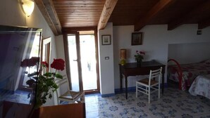 A room with a wooden ceiling, a bed with a floral bedspread, a small table with a chair, and a door leading outside. - Lovers' balcony (Santa Domenica)