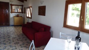 A room with a red sofa, a white table with a bottle and glasses, a wooden cabinet, and a window with a view of greenery. - Lovers' balcony (Santa Domenica)