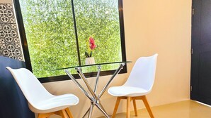 A room with two white chairs and a glass table, a window with a view of greenery, and a tiled floor.
