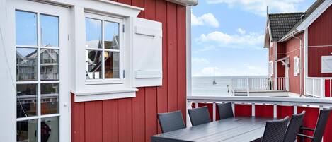 Een rood gebouw met witte accenten, een balkon met een tafel en stoelen, en uitzicht op zee.