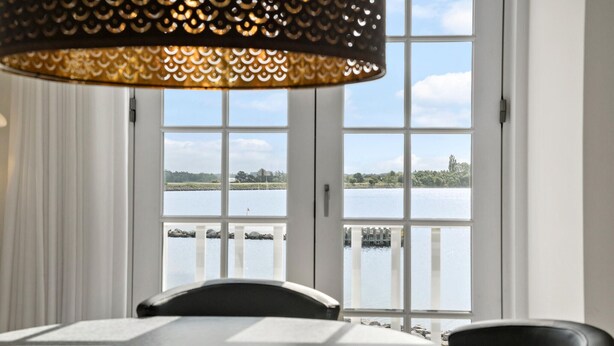 A dining area with a table and chairs, a view of a lake through large windows, and a decorative hanging light fixture.