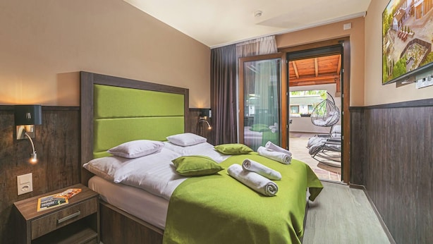 A hotel room with a bed, a nightstand, a window with curtains, and a view of the outside. - Parl Hotel Bükfürdő (Buk)