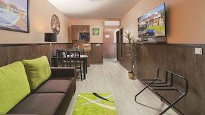 A hotel room with a sofa, a dining table with chairs, a kitchen area, a TV mounted on the wall, and a potted plant. - Parl Hotel Bükfürdő (Buk)