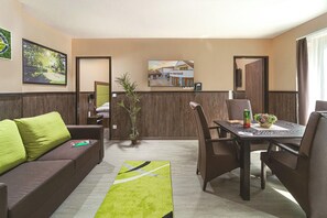 A hotel room with a sofa, dining table, chairs, a plant, a painting, and a TV on the wall. - Parl Hotel Bükfürdő (Buk)