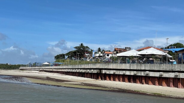 Een kustgebied met een betonnen pier, strand en gebouwen op de achtergrond.