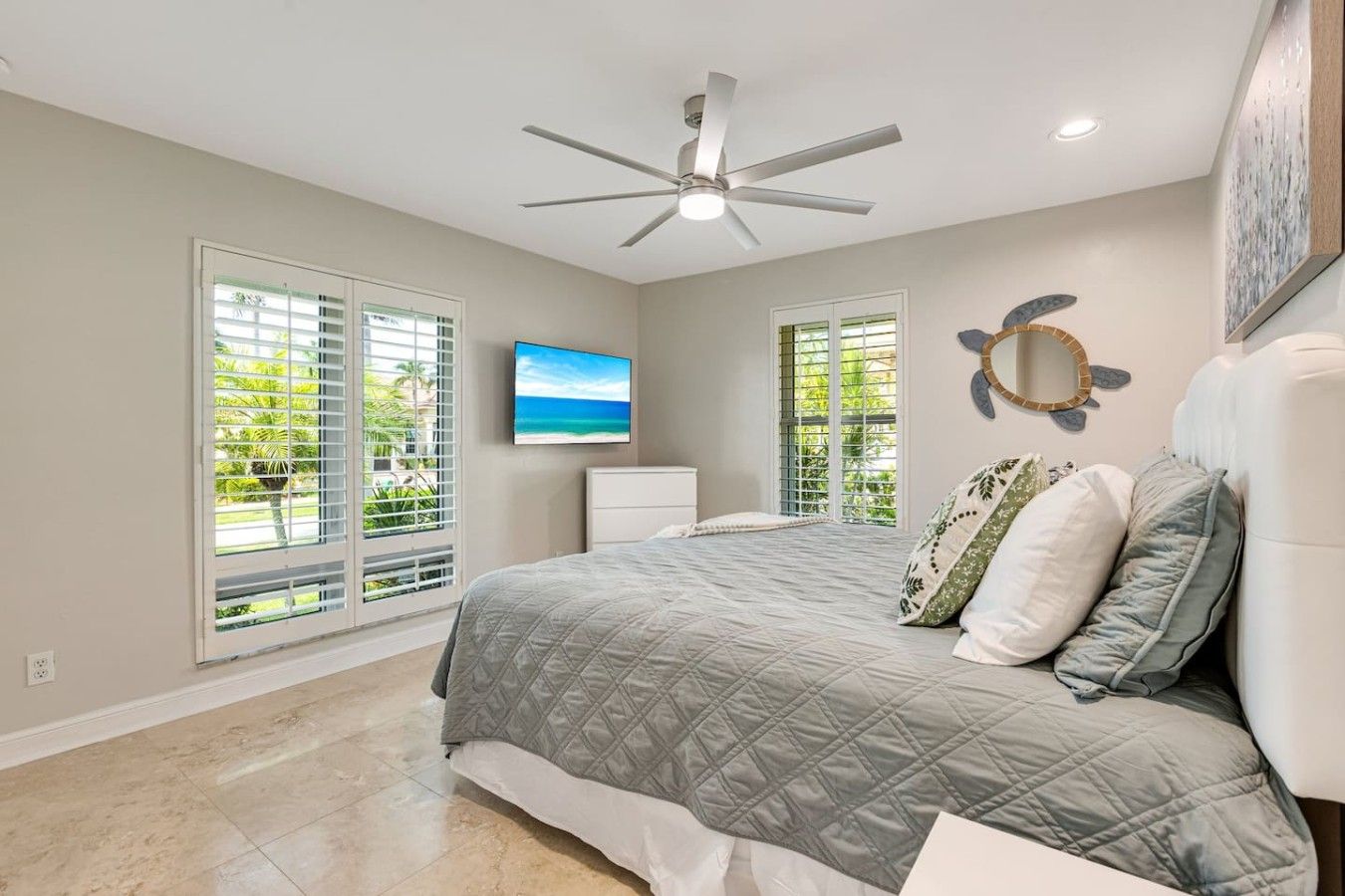 A bedroom with a bed, a ceiling fan, a TV, and large windows with blinds.