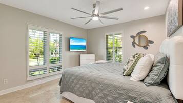 A bedroom with a bed, a ceiling fan, a TV, and large windows with blinds.