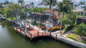 A waterfront property with a dock, a covered patio, and a pool area surrounded by palm trees and greenery.