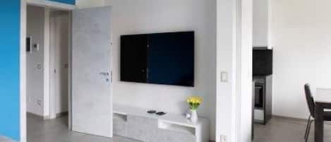 A modern living room with a flat-screen TV mounted on the wall, a white console table with a vase of flowers, and a dining area with a table and chairs.