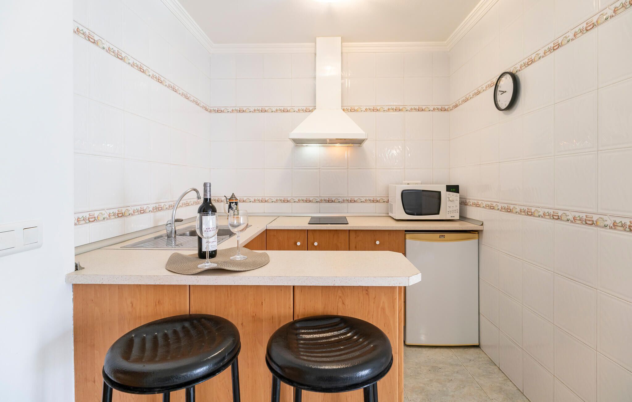 A kitchen with white tiled walls, a white stove, a microwave, a sink, and two stools.