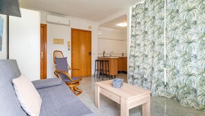 Una sala de estar con un sofá gris, una silla de madera, una mesa de centro de madera y una cocina al fondo.