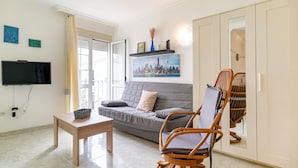 Una sala de estar con un sofá gris, una mesa de centro de madera, un sillón de mimbre, un televisor de pantalla plana y un cuadro con una vista de la ciudad en la pared.