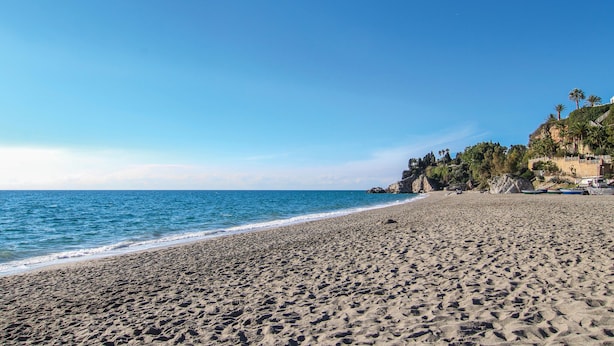 Una playa amplia y arenosa con olas suaves, un cielo azul despejado y un acantilado rocoso con vegetación a la derecha.