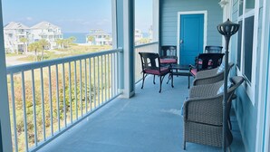 A covered porch with a view of the ocean and houses. There are wicker chairs and a table.