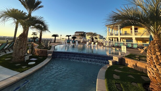 A swimming pool with a waterfall feature, surrounded by palm trees and lounge chairs.