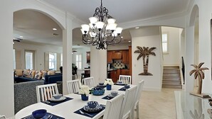A dining area with a white table set for six, surrounded by white chairs, a black chandelier, and a kitchen in the background.