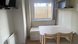Una habitación pequeña y luminosa con suelo de madera, una mesa blanca con dos sillas, un banco, un televisor montado en la pared y una ventana con vistas al exterior.