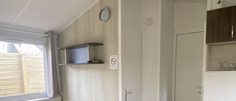 A small, well-lit kitchen with a wooden table and chairs, a window with curtains, a clock, and a no-smoking sign.