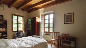 Un dormitorio con una cama, dos sillas, una estantería y ventanas amplias.
