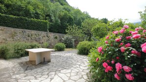 Un patio de piedra con mesa y sillas, rodeado de abundante vegetación y flores rosadas.