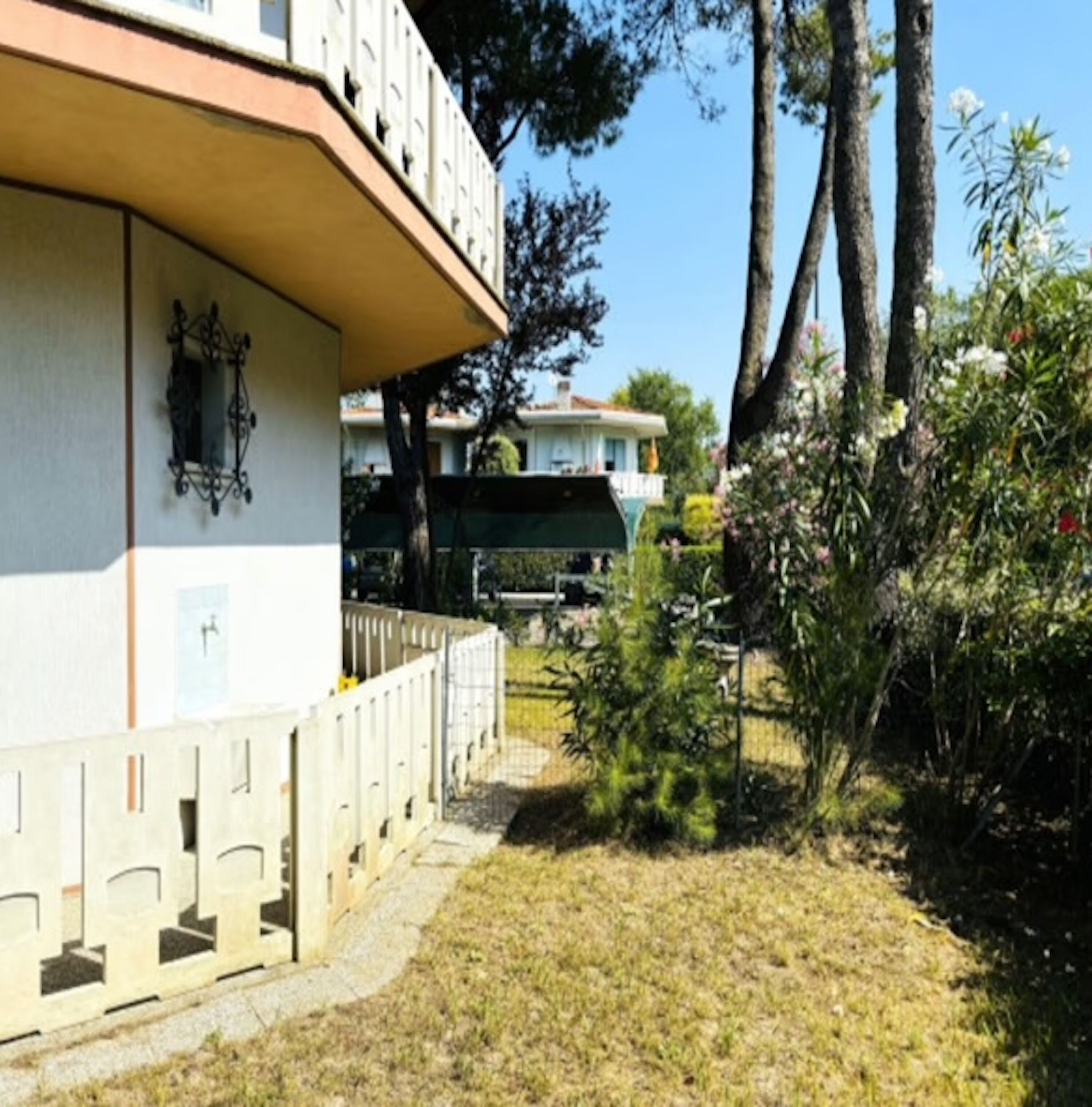 A building with a beige facade, a balcony with a railing, and a small garden area with trees and shrubs.