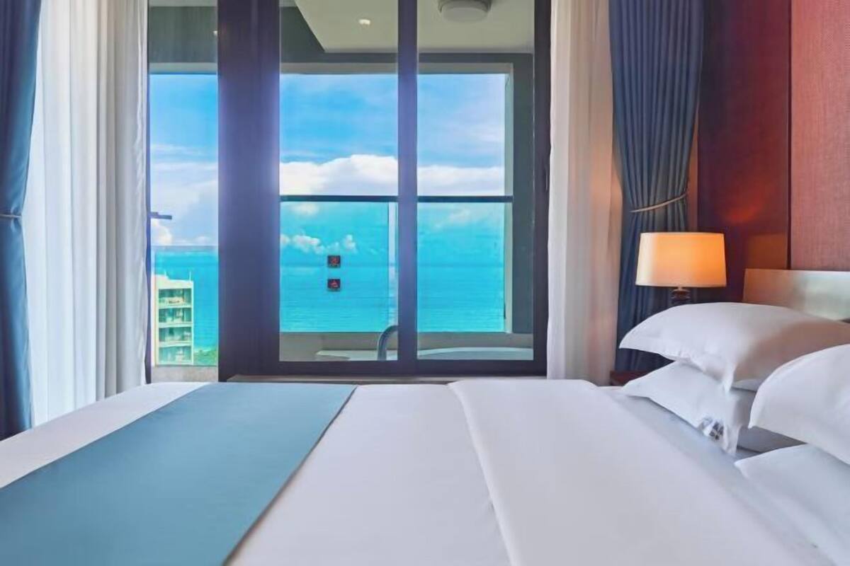 A hotel room with a bed, a nightstand with a lamp, and a view of the ocean through a glass door.