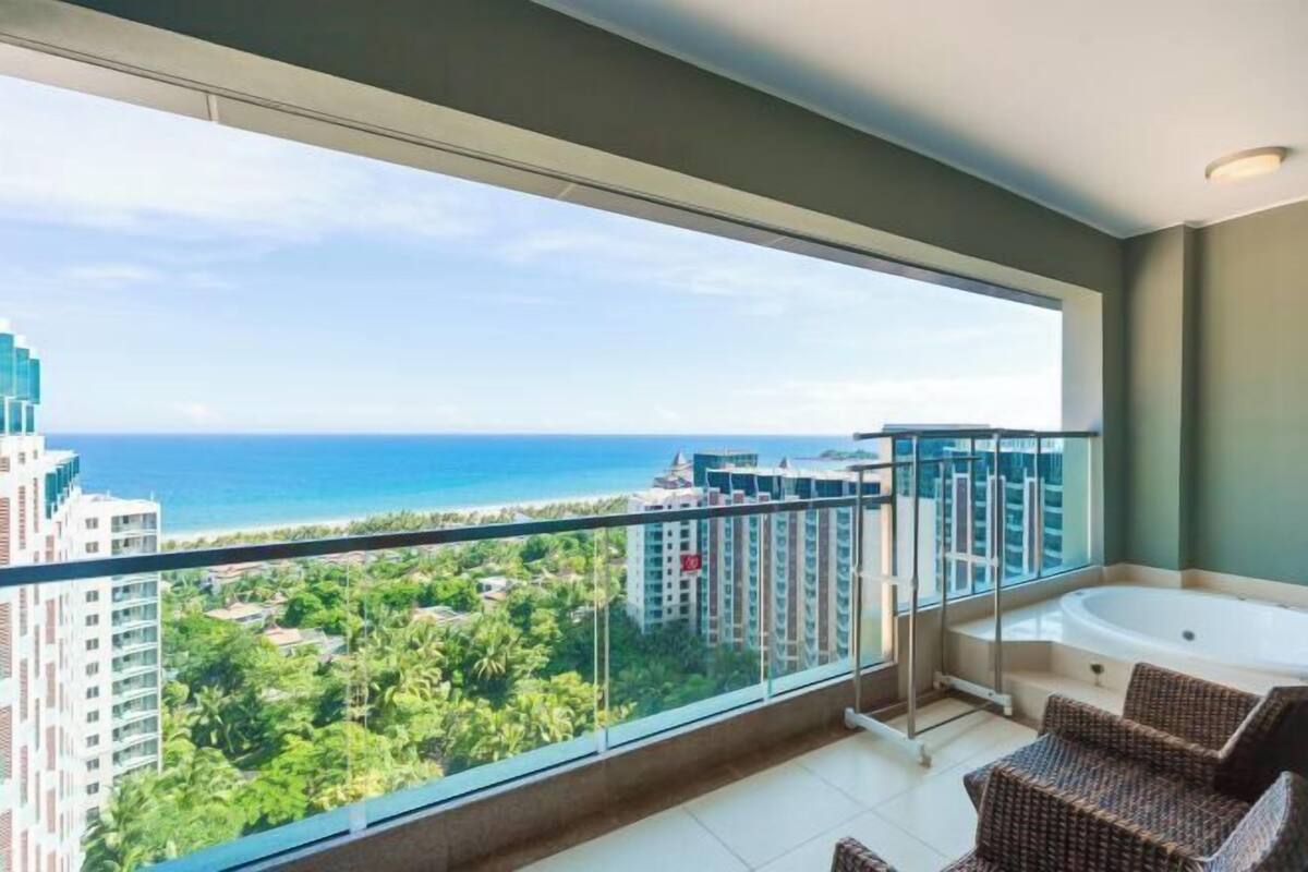 A balcony with a glass railing, a jacuzzi tub, and a view of the ocean and buildings.