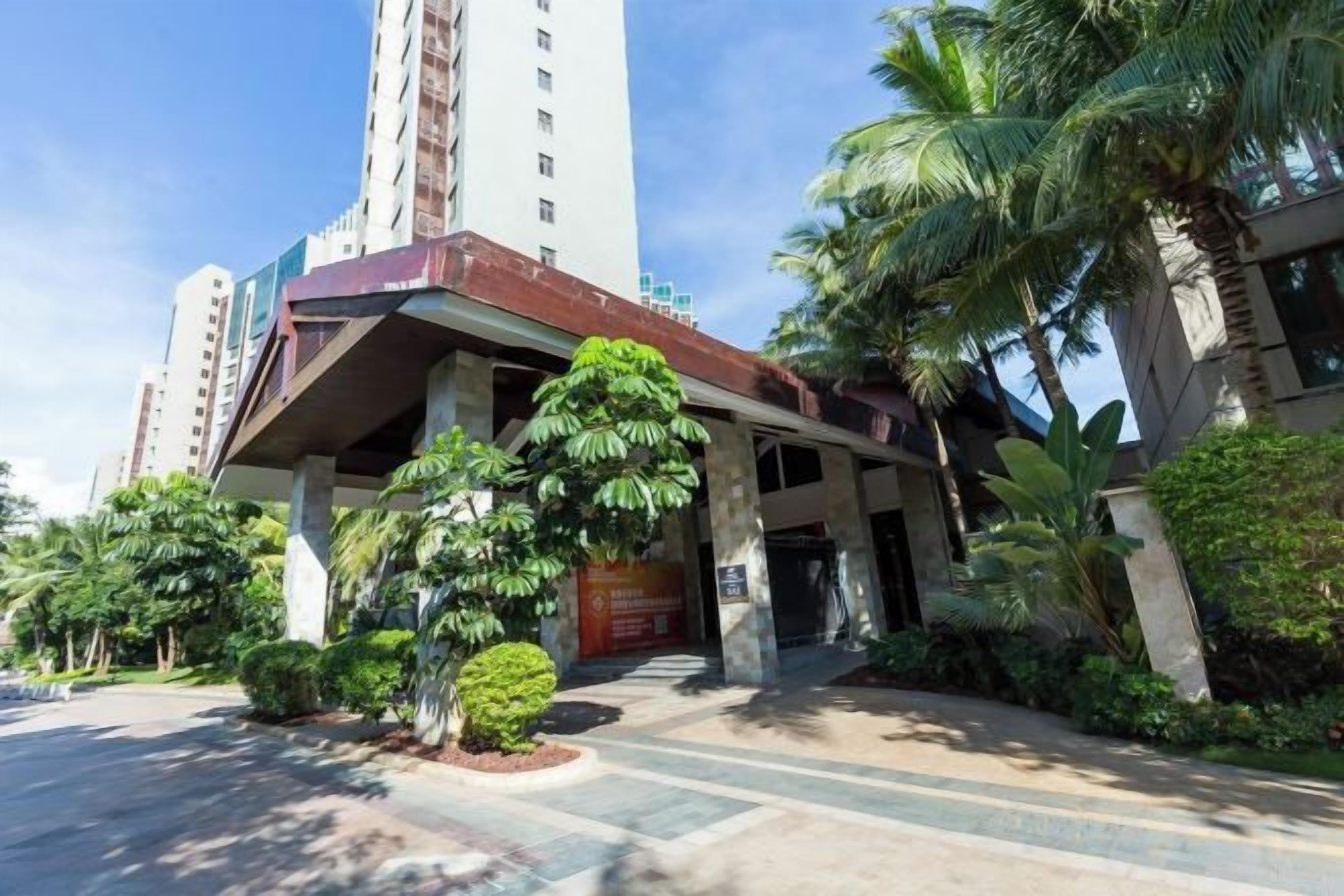 Entrada de un hotel con un corredor techado, palmeras y un edificio alto al fondo.