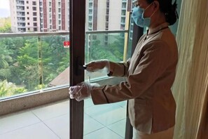 Een persoon met masker en handschoenen, staand bij een raam met uitzicht op groen en gebouwen.